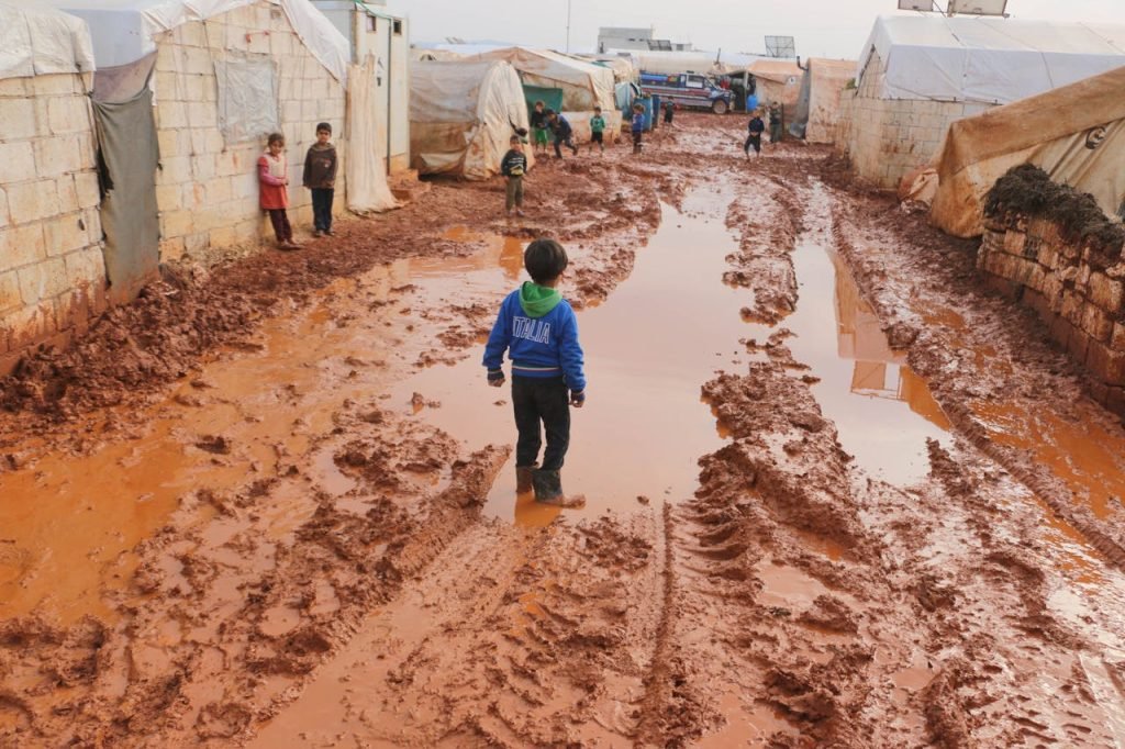 Group of children standing on dirty wet ground with puddles between old tents in refugee camp with in poor settlement